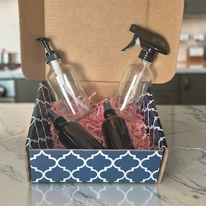 Peut inclure: Une boîte cadeau en carton avec un motif géométrique bleu et blanc contenant deux flacons en verre transparent avec pompes noires, un flacon pulvérisateur en verre transparent avec gâchette noire et deux flacons pulvérisateurs ambrés sur calage rose.