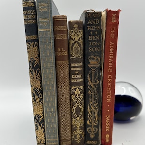 May include: A collection of seven vintage hardcover books with gold lettering and intricate designs on the spines. The books are arranged vertically, with the tallest book on the left and the shortest book on the right. The books are all different colors, including black, brown, red, and blue.