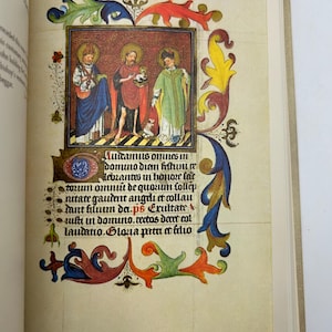 May include: An open book displaying a colorful illuminated manuscript page. The page features a central illustration of three figures in robes, surrounded by ornate floral and foliate borders. Text in Latin is visible.