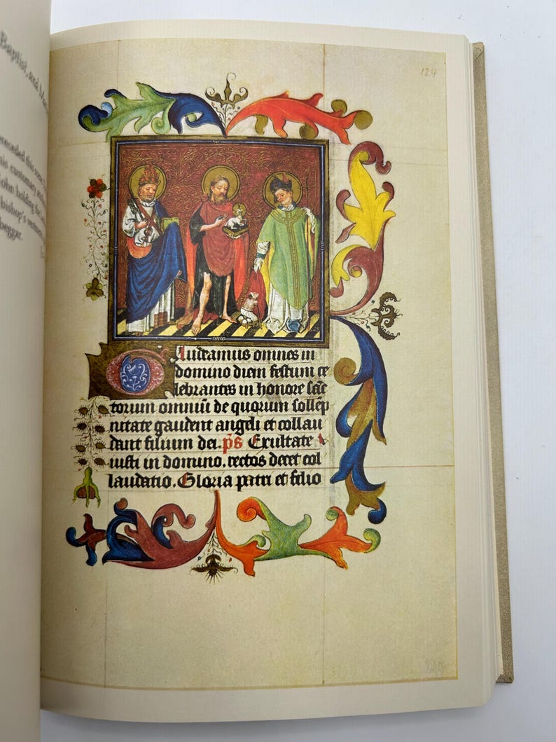 May include: An open book displaying a colorful illuminated manuscript page. The page features a central illustration of three figures in robes, surrounded by ornate floral and foliate borders. Text in Latin is visible.