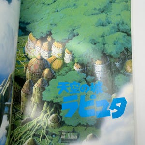 May include: A page from a book featuring a vibrant illustration of a fantasy landscape. The image showcases a floating city with domed structures nestled among lush green foliage. The text "Laputa" is displayed in blue.