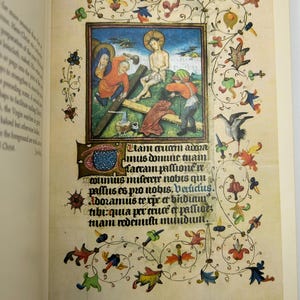 May include: An open book page featuring a medieval illuminated manuscript. The central image depicts a religious scene with figures in colorful robes. Decorative floral and foliate borders frame the illustration, with Latin text below.