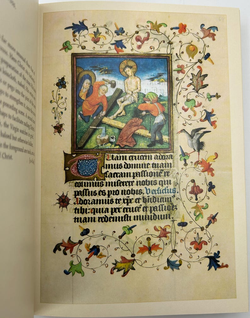 May include: An open book page featuring a medieval illuminated manuscript. The central image depicts a religious scene with figures in colorful robes. Decorative floral and foliate borders frame the illustration, with Latin text below.