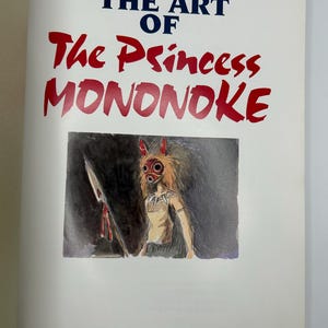 May include: A book cover titled THE ART OF The Princess MONONOKE in dark blue and red text. Below the title is a watercolor illustration of a figure wearing a red and white mask with pointed ears, holding a spear against a dark gray background.