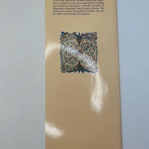 Op de afbeelding: De achterkant van een boek met een gedetailleerde illustratie van een middeleeuws knoopontwerp in goud en rood. De tekst op de achterkant beschrijft de expertise van de auteur in middeleeuwse manuscripten en zijn werk bij Sotheby's.
