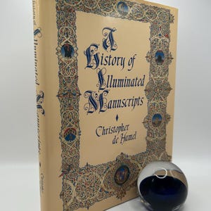Op de afbeelding: Een boek getiteld "A History of Illuminated Manuscripts" van Christopher de Hamel. De omslag heeft een sierlijk, kleurrijk randontwerp met een middeleeuws lettertype.