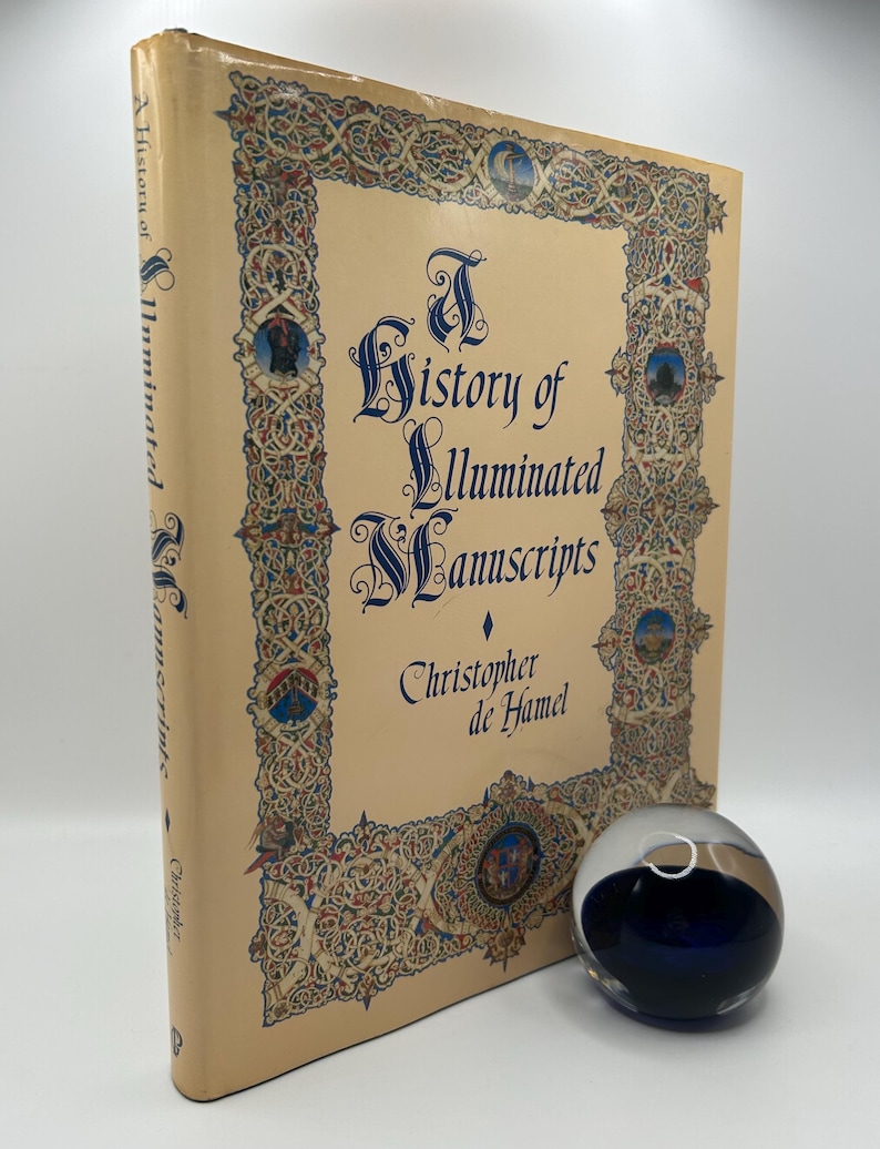 Op de afbeelding: Een boek getiteld "A History of Illuminated Manuscripts" van Christopher de Hamel. De omslag heeft een sierlijk, kleurrijk randontwerp met een middeleeuws lettertype.
