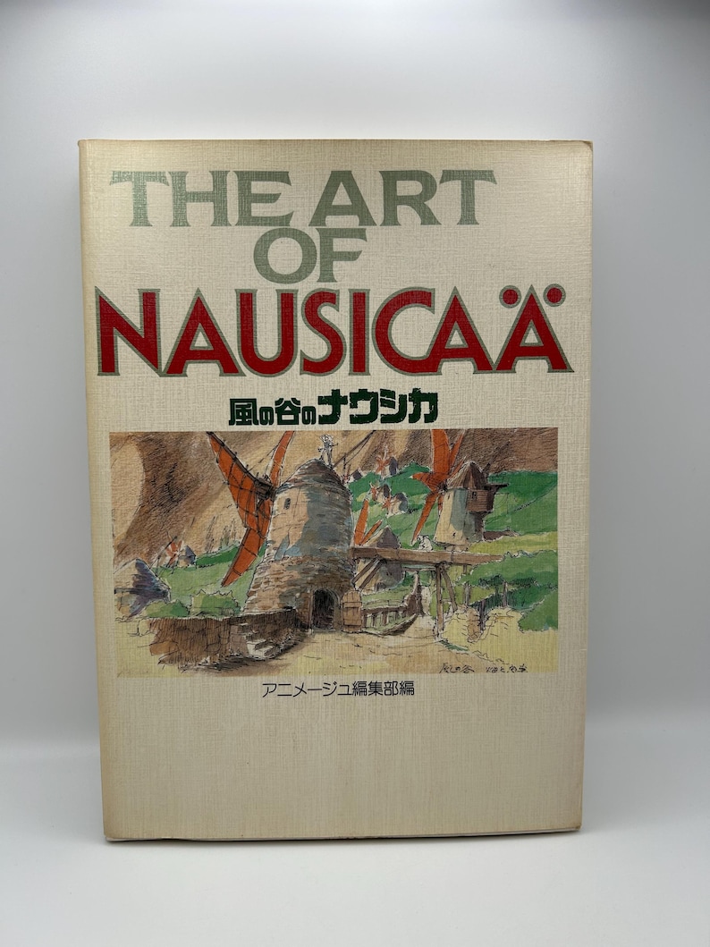 May include: A book titled "THE ART OF NAUSICA&Auml;" with Japanese text and an illustration of windmills. The cover is beige with red and green text. The illustration depicts a rural landscape with windmills.