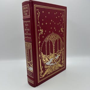 Op de afbeelding: Een hardcover boek getiteld "Tales of Mystery and Imagination" van Edgar Allan Poe. Het boek heeft een dieprode kaft met gouden accenten, waaronder sterren en een sierlijke illustratie. De rug heeft ook gouden letters.