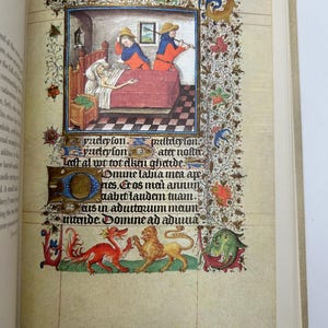 May include: An open book page featuring a medieval illustration. The artwork depicts a scene with figures in period clothing, a bed, and decorative borders. The page also includes handwritten text and ornate embellishments.