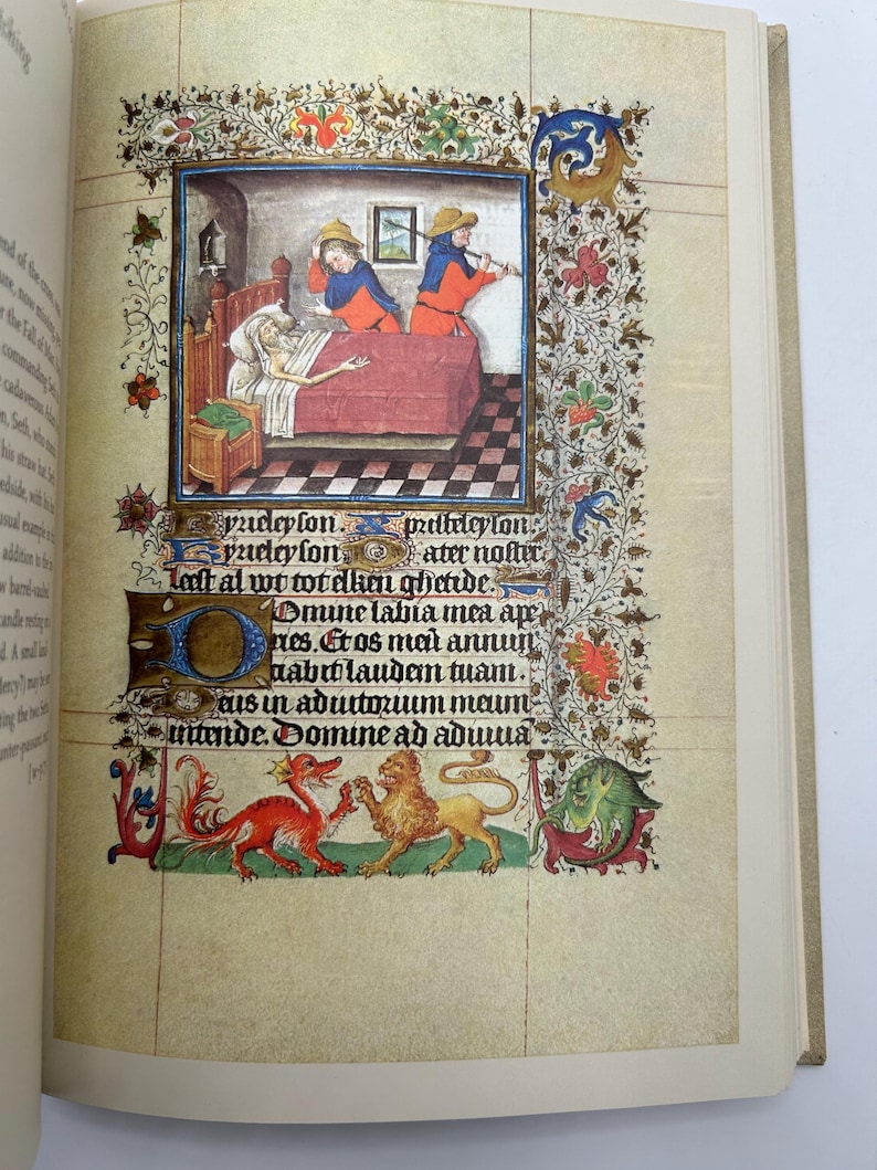 May include: An open book page featuring a medieval illustration. The artwork depicts a scene with figures in period clothing, a bed, and decorative borders. The page also includes handwritten text and ornate embellishments.