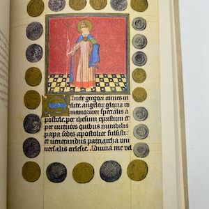 May include: Open book page featuring a medieval illustration. A central image depicts a robed figure with a halo, holding a staff, against a red backdrop. The page is bordered by gold and silver coin illustrations and includes handwritten text.