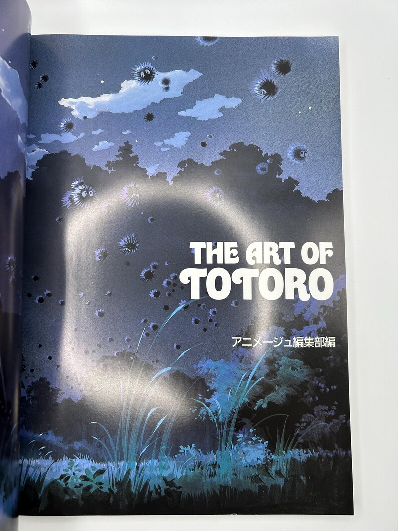 Puede incluir: Portada del libro "THE ART OF TOTORO" con letras blancas. La ilustraci&oacute;n muestra una escena nocturna de bosque en tonos azul oscuro y verde, con duendes de holl&iacute;n flotantes y texto japon&eacute;s.