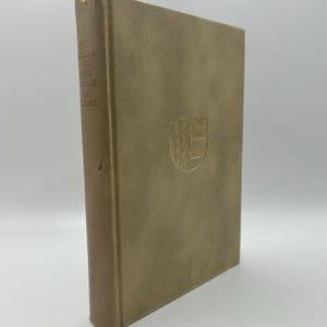 May include: A hardcover book with a tan cover and embossed text and emblem. The book's spine displays the title "THE SOUPS OF EARTH-ERGEVE OF LEVES". A gold emblem is centered on the front cover.