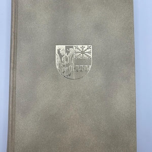 May include: A hardcover book with a textured, light gray cover. A silver emblem is centered on the front, featuring a detailed design of a shield with various elements. The spine is a slightly darker shade of gray.