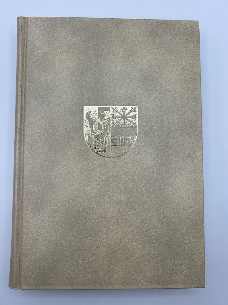 May include: A hardcover book with a textured, light gray cover. A silver emblem is centered on the front, featuring a detailed design of a shield with various elements. The spine is a slightly darker shade of gray.