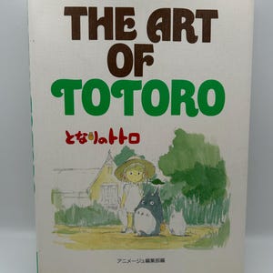 Puede incluir: Un libro titulado "The Art of Totoro" con texto verde y marr&oacute;n. La portada presenta una ilustraci&oacute;n de una ni&ntilde;a con Totoro y una peque&ntilde;a criatura blanca en un campo. Tambi&eacute;n hay texto e ilustraciones japonesas.