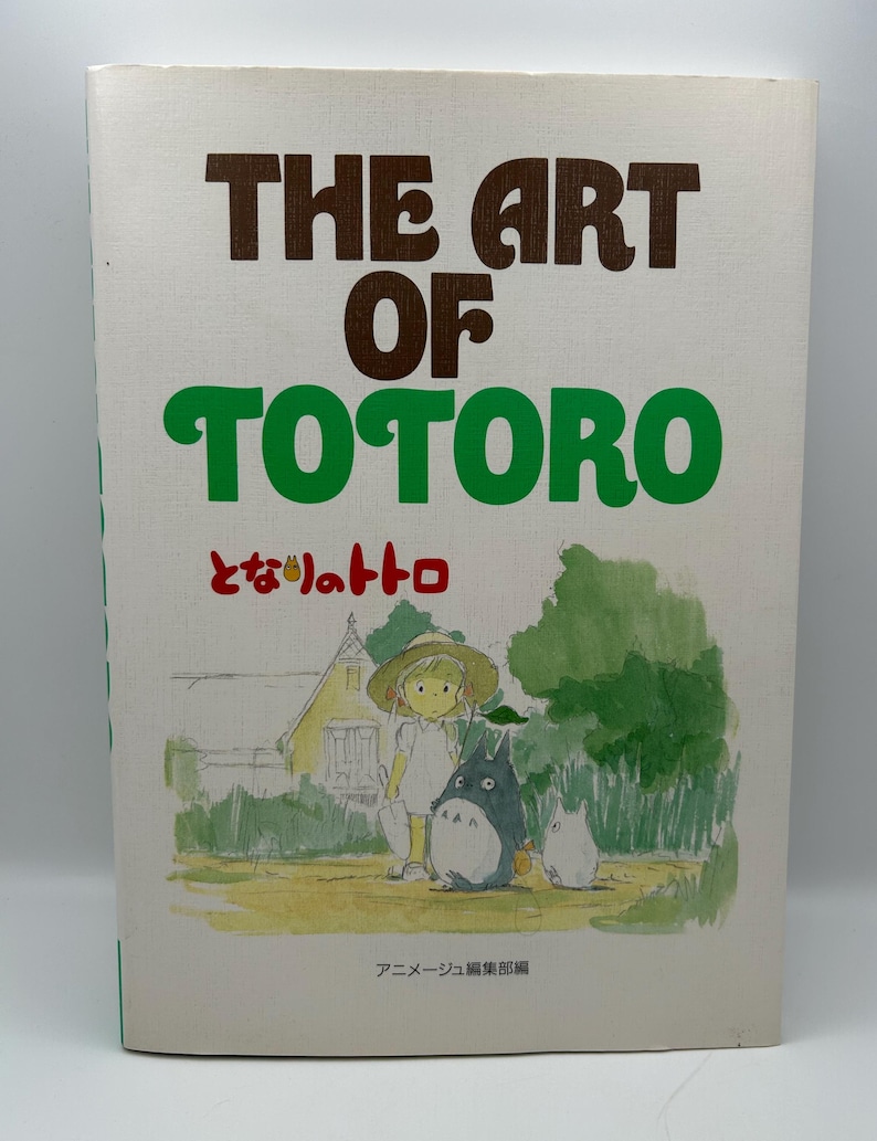 Puede incluir: Un libro titulado "The Art of Totoro" con texto verde y marr&oacute;n. La portada presenta una ilustraci&oacute;n de una ni&ntilde;a con Totoro y una peque&ntilde;a criatura blanca en un campo. Tambi&eacute;n hay texto e ilustraciones japonesas.