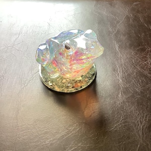 May include: Iridescent glass frog figurine with a textured surface. The frog sits on a clear, circular base. The glass reflects a rainbow of colors, including pink, yellow, and blue.