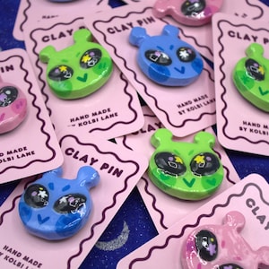 Pode incluir: Uma coleção de pins de argila feitos à mão em várias cores, incluindo azul, verde e rosa. Cada pin apresenta um design estilizado de rosto alienígena com olhos grandes e uma boca pequena. Os pins são exibidos em cartões cor-de-rosa com o texto "CLAY PIN" e "HAND MADE BY KOLBI LANE."