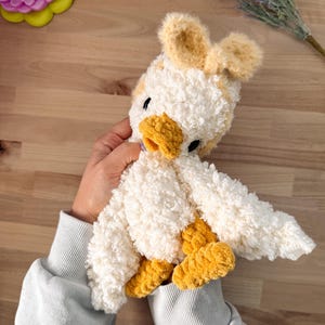 May include: A handmade plush duck toy with white, yellow, and tan accents. The duck has a fluffy, textured body, yellow feet and beak, and small black eyes. The duck is being held by a person wearing a light blue sweatshirt.