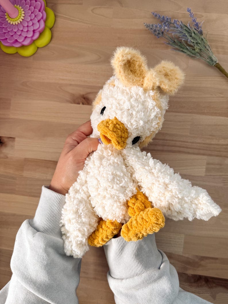 May include: A handmade plush duck toy with white, yellow, and tan accents. The duck has a fluffy, textured body, yellow feet and beak, and small black eyes. The duck is being held by a person wearing a light blue sweatshirt.