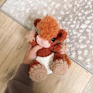 Puede incluir: Un peluche de vaca hecho a mano con ganchillo en tonos marr&oacute;n, beige y blanco. El peluche tiene un aspecto esponjoso y texturizado con una nariz rosa claro y ojos negros. La vaca lleva un pa&ntilde;al blanco.