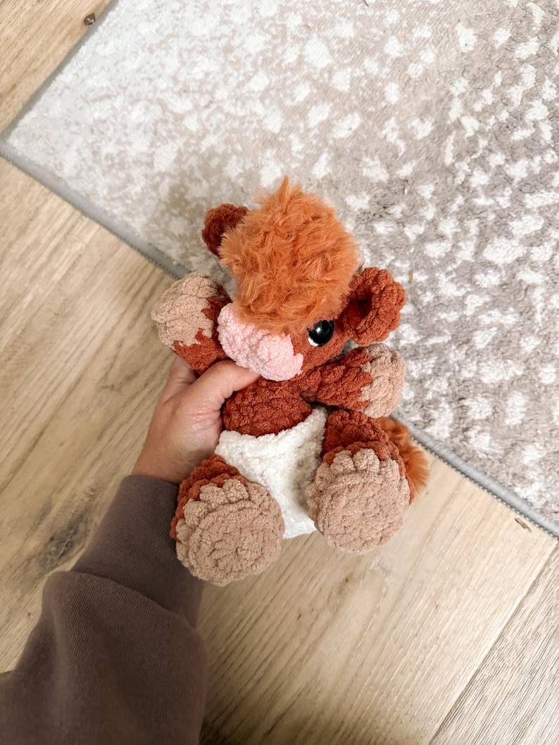 Puede incluir: Un peluche de vaca hecho a mano con ganchillo en tonos marr&oacute;n, beige y blanco. El peluche tiene un aspecto esponjoso y texturizado con una nariz rosa claro y ojos negros. La vaca lleva un pa&ntilde;al blanco.