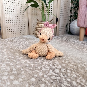 May include: A handmade, beige crochet duck with peach-colored accents on its beak, feet, and wings. The duck is wearing a pink bow on its head. The plush toy is sitting on a patterned rug.