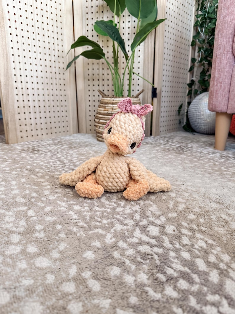 May include: A handmade, beige crochet duck with peach-colored accents on its beak, feet, and wings. The duck is wearing a pink bow on its head. The plush toy is sitting on a patterned rug.