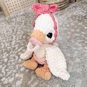 May include: A handmade crochet duckling plushie. The duckling is primarily white with a pink bow, beak, and accents. It has black safety eyes and orange feet. The plushie is sitting on a textured surface.