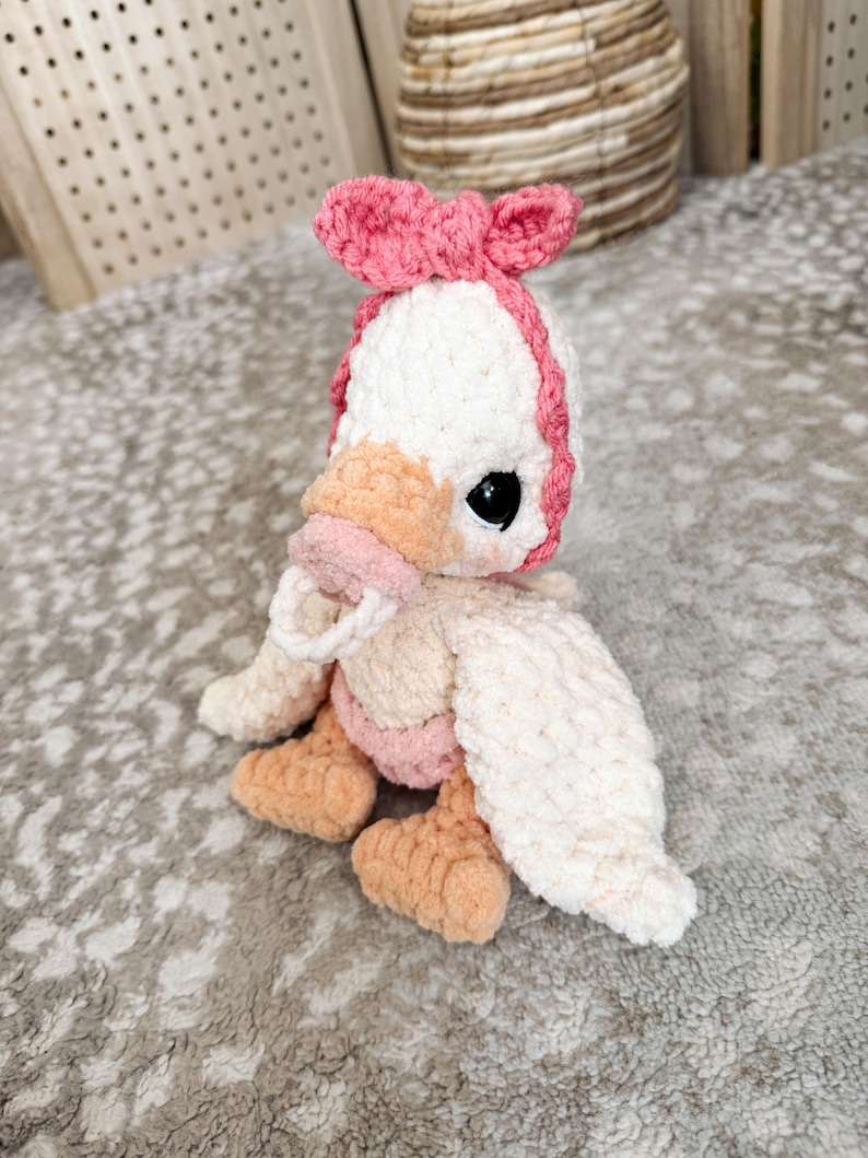 May include: A handmade crochet duckling plushie. The duckling is primarily white with a pink bow, beak, and accents. It has black safety eyes and orange feet. The plushie is sitting on a textured surface.