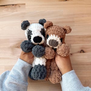 Peut inclure: Deux ours en peluche crochet&eacute;s &agrave; la main. L'un est blanc avec des accents noirs, l'autre est marron avec un museau blanc. Les deux ont des yeux fonc&eacute;s et des corps duveteux et textur&eacute;s. Les ours sont tenus devant un fond en bois.