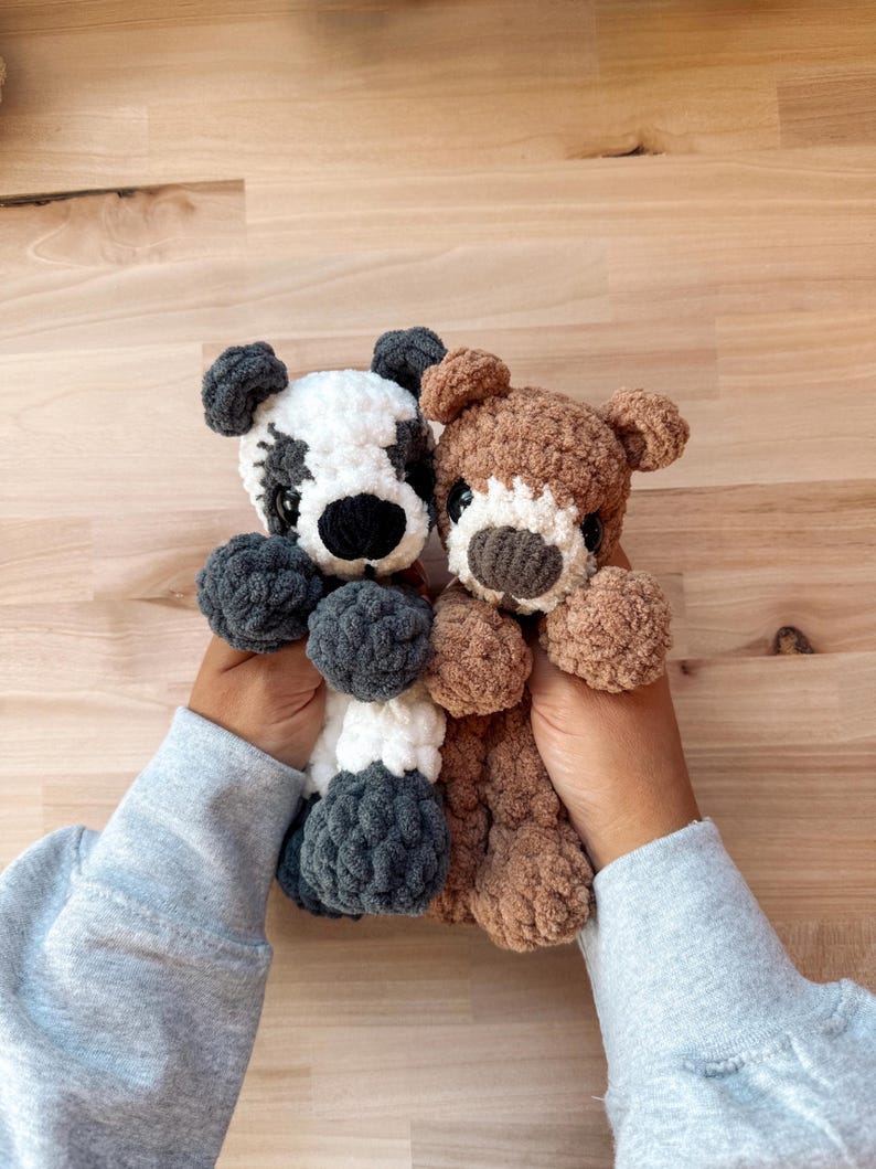 Peut inclure: Deux ours en peluche crochet&eacute;s &agrave; la main. L'un est blanc avec des accents noirs, l'autre est marron avec un museau blanc. Les deux ont des yeux fonc&eacute;s et des corps duveteux et textur&eacute;s. Les ours sont tenus devant un fond en bois.