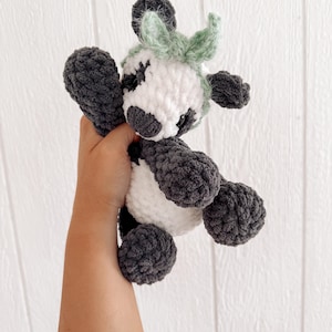 May include: A handmade crochet cow plushie in white, gray, and light green. The plush toy has a rounded body, with gray legs, arms, and spots around the eyes. The cow has a white face and a light green tuft of hair.