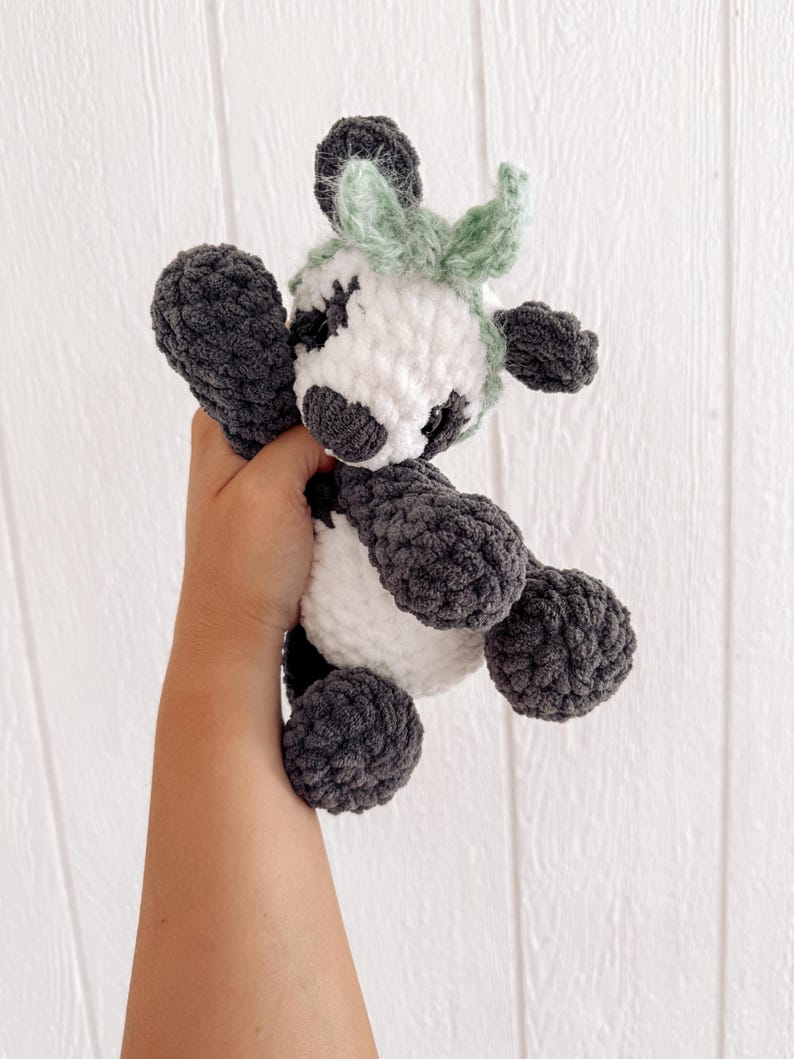 May include: A handmade crochet cow plushie in white, gray, and light green. The plush toy has a rounded body, with gray legs, arms, and spots around the eyes. The cow has a white face and a light green tuft of hair.