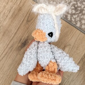 May include: A handmade, plush duckling toy in soft blue, peach, and white. The duckling has fluffy wings, a tuft of white hair, and black eyes. The feet and beak are a warm peach color.