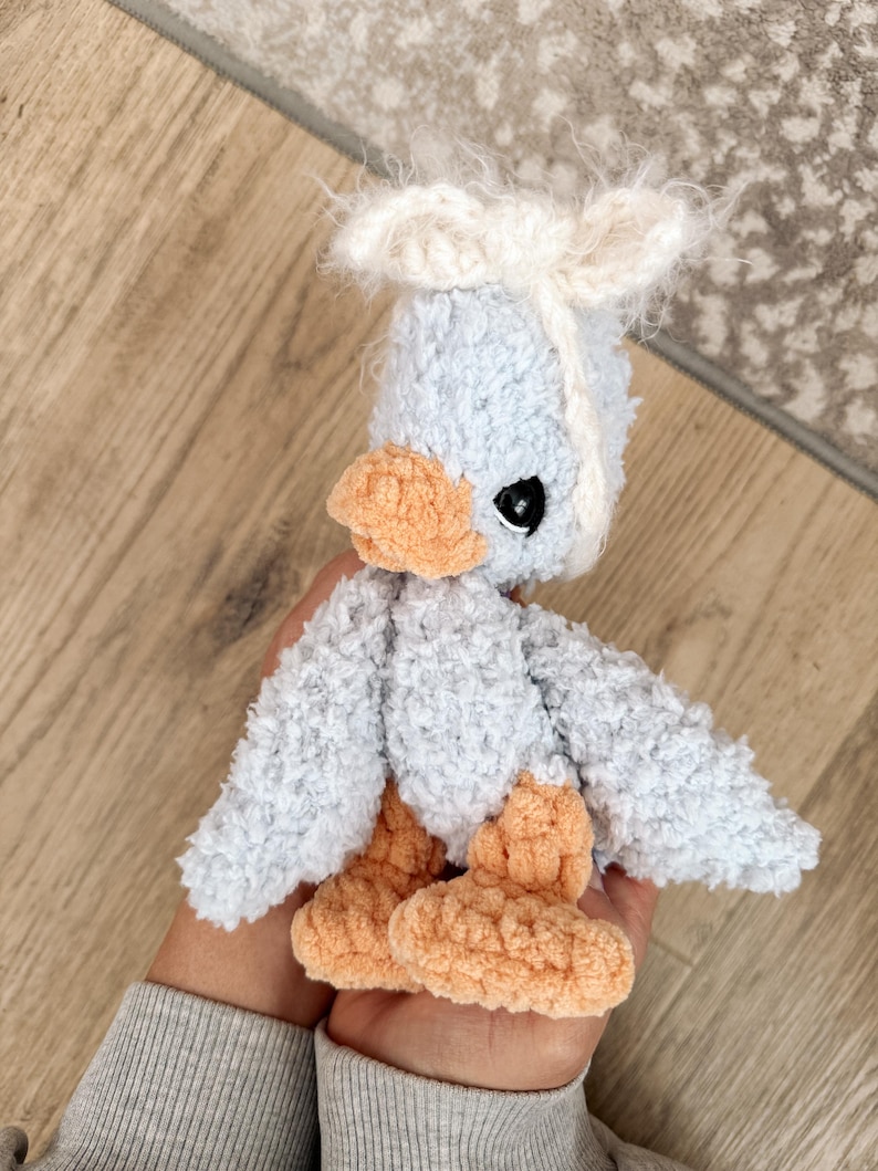 May include: A handmade, plush duckling toy in soft blue, peach, and white. The duckling has fluffy wings, a tuft of white hair, and black eyes. The feet and beak are a warm peach color.
