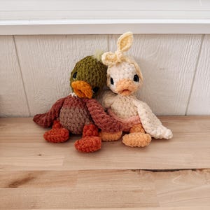 May include: Two handmade crochet duck plushies. One duck is green, brown, and orange, the other is cream, peach, and yellow. Both have black eyes and orange beaks. The duck on the right has bunny ears.