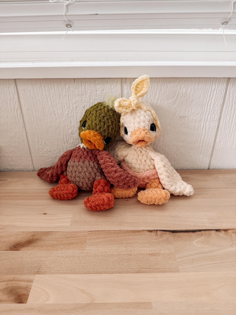 May include: Two handmade crochet duck plushies. One duck is green, brown, and orange, the other is cream, peach, and yellow. Both have black eyes and orange beaks. The duck on the right has bunny ears.