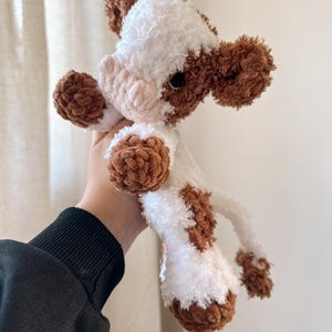 May include: A crocheted plush toy cow with brown and white fur. The cow has a pink nose and black eyes.