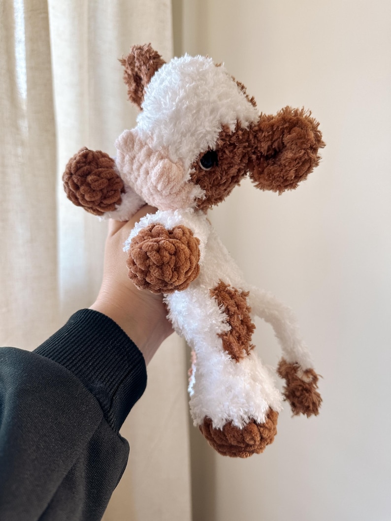 May include: A crocheted plush toy cow with brown and white fur. The cow has a pink nose and black eyes.