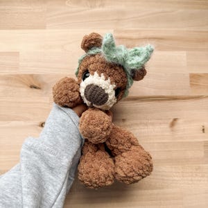 Peut inclure: Un ours en peluche marron fait &agrave; la main avec un museau blanc et un nez marron fonc&eacute;. L'ours a un n&oelig;ud vert clair sur la t&ecirc;te. L'ours est fait d'une mati&egrave;re douce et textur&eacute;e.