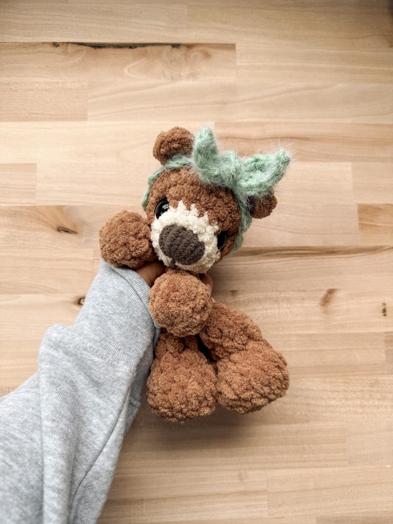 Peut inclure: Un ours en peluche marron fait &agrave; la main avec un museau blanc et un nez marron fonc&eacute;. L'ours a un n&oelig;ud vert clair sur la t&ecirc;te. L'ours est fait d'une mati&egrave;re douce et textur&eacute;e.