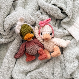 May include: Two handmade crochet duck toys. One duck is wearing a green and white hat, and the other is wearing a pink bow. The ducks have orange beaks and are resting on a soft, light green blanket.