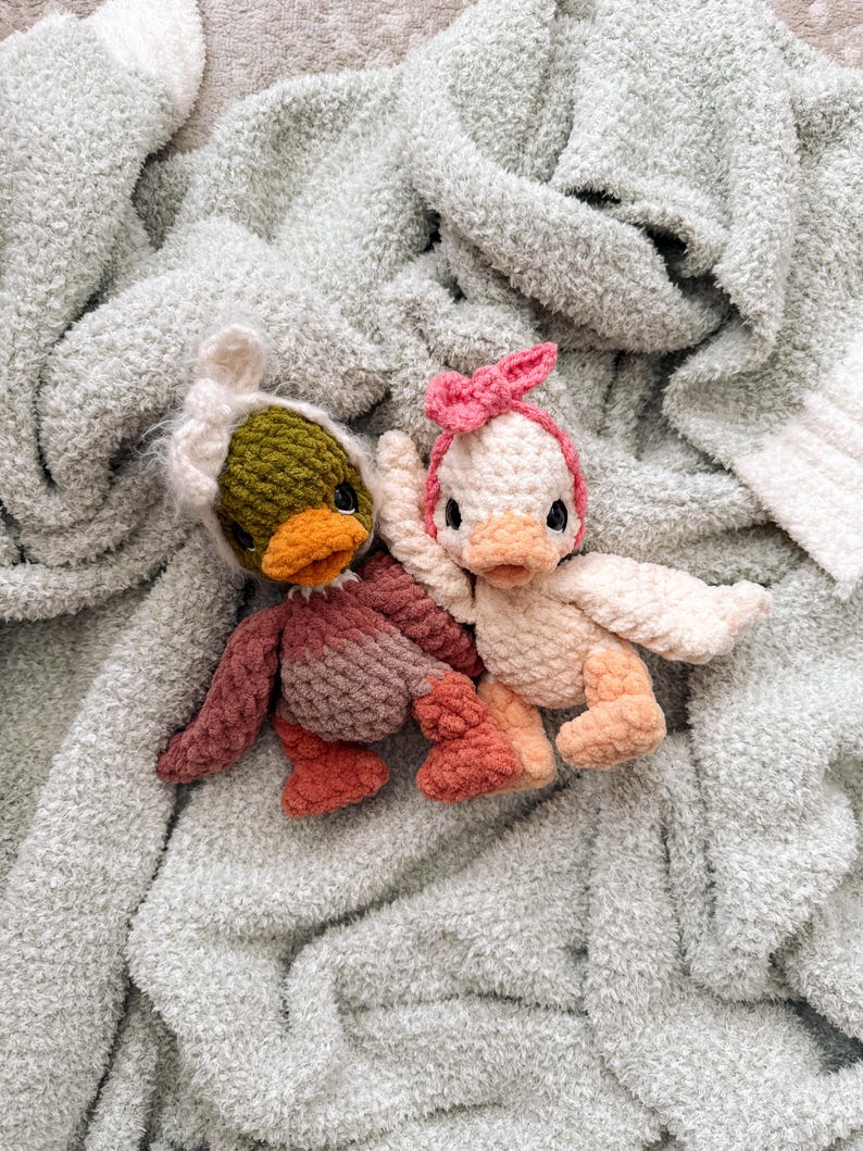 May include: Two handmade crochet duck toys. One duck is wearing a green and white hat, and the other is wearing a pink bow. The ducks have orange beaks and are resting on a soft, light green blanket.