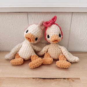 May include: Two handmade crochet duck dolls. One duck has a pink bow and the other has a pink head covering. Both ducks have white bodies, orange beaks and feet, and black eyes. The dolls are sitting on a light brown wooden surface.