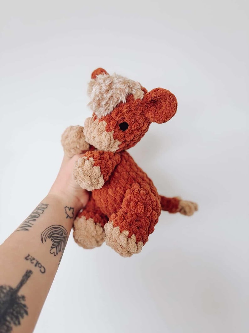 Puede incluir: Un peluche de caballo de ganchillo hecho a mano en tonos &oacute;xido y beige. El animal de peluche tiene un aspecto texturizado y suave con una melena y pezu&ntilde;as de color marr&oacute;n claro. El juguete se sostiene sobre un fondo blanco.