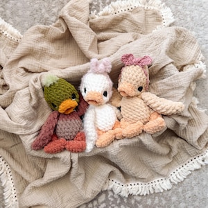 May include: Three handmade crochet duck toys in shades of green, white, and peach, each with a unique hat or bow. The ducks are resting on a beige, fringed blanket, creating a cozy and whimsical scene.