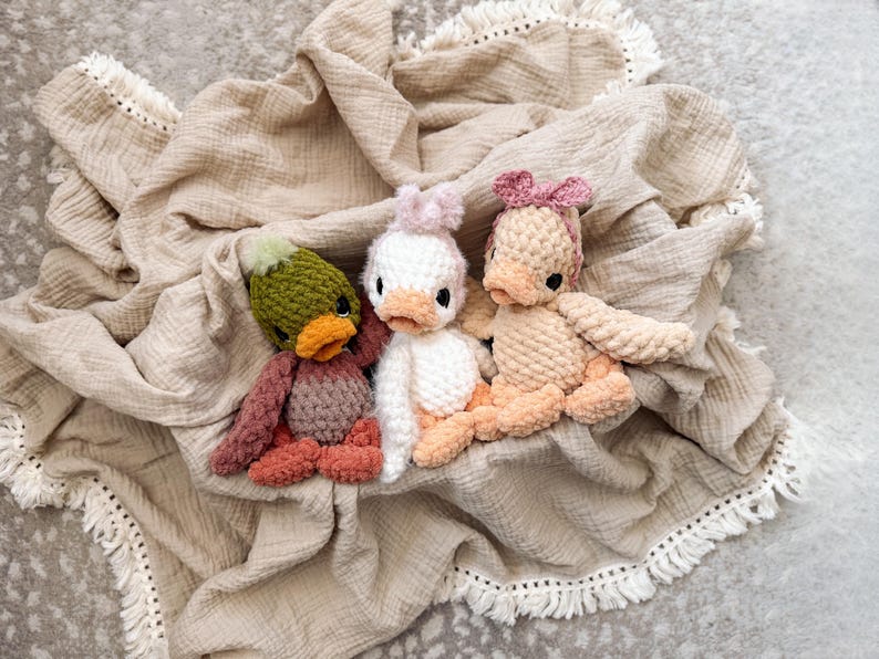 May include: Three handmade crochet duck toys in shades of green, white, and peach, each with a unique hat or bow. The ducks are resting on a beige, fringed blanket, creating a cozy and whimsical scene.